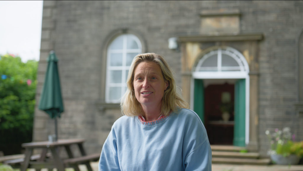 Andrea from Wainsgate Chapel smiling outside the historic stone building during her case study interview.