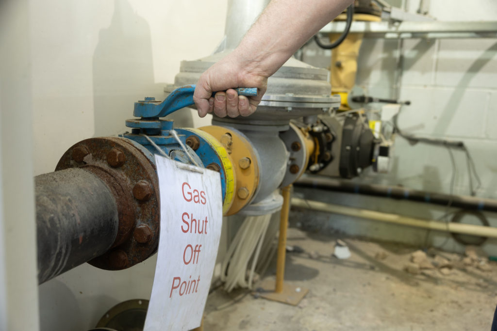 Close-up of an engineer's hand turning off the yellow master commercial gas isolation valve inside the old plant room.